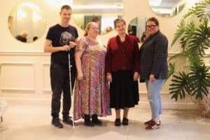 Cztery osoby stoją w eleganckim wnętrzu z lustrami na ścianie. Od lewej: mężczyzna w ciemnej koszulce z białą laską, kobieta w kolorowej sukience, kobieta w bordowym żakiecie i czarnej spódnicy oraz kobieta w ciemnym żakiecie, okularach i dżinsach.