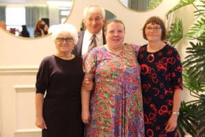 Cztery uśmiechnięte osoby stoją na tle ściany z okrągłymi lustrami. Z przodu są trzy kobiety: po bokach dwie w ciemnych sukienkach, a pośrodku kobieta w kolorowej sukience. Za nimi stoi mężczyzna w garniturze.