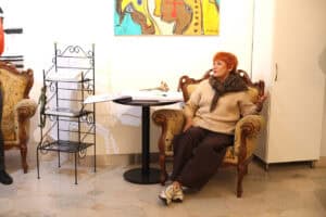 Maria Pietrusza-Budzyńska siedzi w ozdobnym fotelu i gestykuluje podczas rozmowy. Obok stoją mały stolik i metalowy stojak z materiałami promocyjnymi.