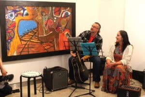 Mężczyzna z gitarą akustyczną i uśmiechnięta kobieta w barwnej sukni oraz białym swetrze siedzą podczas występu muzycznego w galerii. Za nimi wisi duży obraz przedstawiający tory kolejowe. Obok znajdują się sprzęt nagłośnieniowy i stołek z napisem „Teatroterapia Lubelska”.