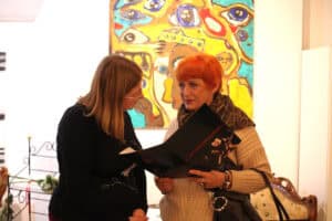 Maria Pietrusza-Budzyńska i druga kobieta wspólnie przeglądają otwarty folder. Maria trzyma go w dłoniach, a druga kobieta przygląda się jego zawartości. Na ścianie za nimi wisi barwny obraz z motywem oczu.
