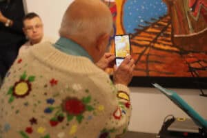 Mężczyzna w jasnym swetrze we wzory kwiatowe fotografuje smartfonem obraz przedstawiający tory kolejowe.