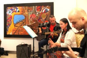 Kobieta śpiewa do mikrofonu, a obok niej mężczyzna gra na gitarze akustycznej. Drugi gitarzysta również uczestniczy w występie. Za muzykami wisi obraz.