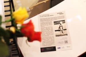 Otwarta księga pamiątkowa lub wydruk opisu wystawy „Co się z nami stało. Wojna...” Marcina Wrzala leży obok żółtych i czerwonych róż.