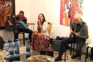 Wokalistka w kolorowej sukni śpiewa do mikrofonu, a dwóch gitarzystów akompaniuje jej podczas koncertu na wernisażu. Na ścianach galerii wiszą barwne obrazy.