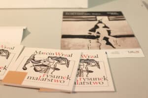 Na jasnym blacie leżą katalogi z napisem „Marcin Wrzal – rysunek, malarstwo” oraz czarno-biały plakat wystawy z logotypami partnerów, w tym Miasta Lublin.