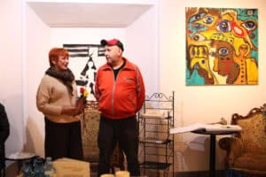 Maria Pietrusza-Budzyńska trzyma kilka róż i rozmawia z Marcinem Wrzalem. Stoją w galerii, na której ścianach wiszą kolorowe obrazy abstrakcyjne oraz czarno-biała grafika.