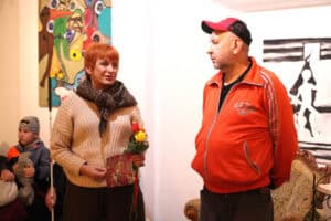 Maria Pietrusza-Budzyńska trzyma róże i plakat wystawy, a obok niej stoi mężczyzna w pomarańczowej bluzie i czapce z daszkiem. Rozmawiają w galerii, gdzie na ścianie wisi kolorowy obraz, a goście wernisażu siedzą w pobliżu.