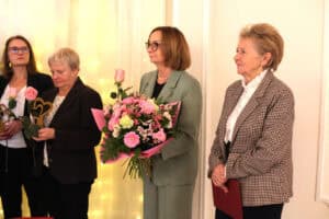 Cztery kobiety w eleganckich strojach stoją w rzędzie. W dłoniach trzymają bukiety kwiatów i pamiątkowe statuetki w kształcie serc.