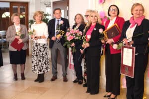 Grupa gości stoi w rzędzie na tle dekoracji ze światłami. W rękach trzymają kwiaty, statuetki, dyplomy i wyróżnienia.