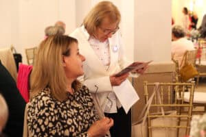 Kobieta w białym garniturze czyta program lub broszurę i rozmawia z kobietą obok niej. W sali stoją złote krzesła, a wokół są goście.