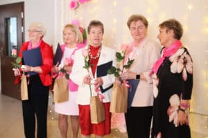 Grupa kobiet w eleganckich strojach stoi w rzędzie. W dłoniach trzymają pamiątkowe dyplomy, teczki, różowe róże i małe torby z prezentami.