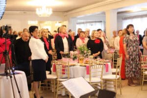 Uczestnicy spotkania stoją na sali bankietowej. W pierwszym rzędzie są między innymi kobieta w białym żakiecie i ksiądz w sutannie.