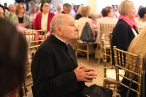Ksiądz w sutannie siedzi przy stole wśród uczestników uroczystości. Ma złożone ręce.