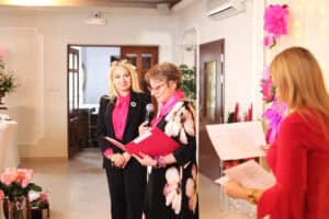 Grażyna Chaberko w czarnej sukni w kwiaty przemawia do gości przez mikrofon. Obok niej stoi Anna Augustyniak.