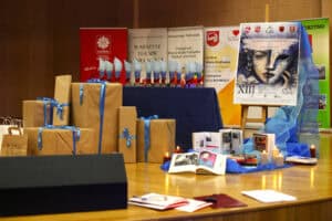 Stół konferencyjny na scenie. Obok stoją paczki różnej wielkości w brązowym papierze z niebieskimi wstążkami. Na stole w rzędach stoją błękitne statuetki, książki i płonące świece. W tle banery i plakat festiwalu.