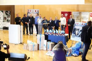 Grupa osób na scenie podczas uroczystości wręczenia nagród. Niektóre osoby trzymają dyplomy i upominki. W tle jasna ściana z drewnianych paneli.