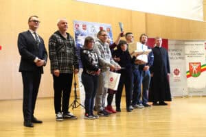 Uśmiechnięty laureat konkursu unosi statuetkę i dyplom na scenie. Obok stoją organizatorzy i inni uczestnicy.
