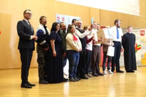 Grupa kilkunastu osób na scenie odbiera nagrody, statuetki i paczki z upominkami. Towarzyszą im oficjalni goście.