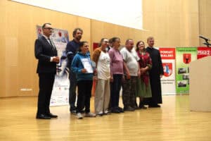 Uczestniczka wydarzenia trzyma dyplom i statuetkę. Obok niej stoi grupa laureatów na scenie. W tle ściana z drewnianych paneli.