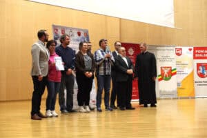 Grupa laureatów i uczestników festiwalu pozuje na scenie z księdzem. Jedna osoba trzyma dyplom, a inna statuetkę.