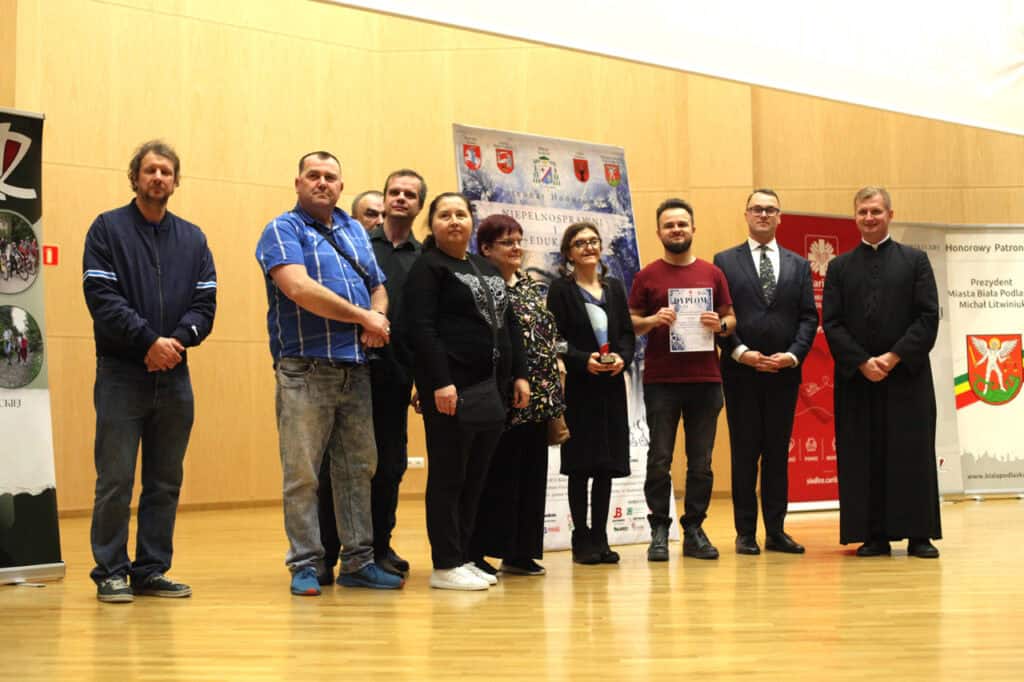 Grupa osób na scenie podczas ceremonii wręczenia nagród. Wśród nich ksiądz w sutannie i mężczyzna w garniturze.