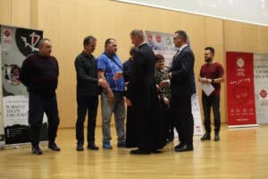 Ksiądz, mężczyzna w garniturze i kilka innych osób witają uczestników na scenie uściskiem dłoni.