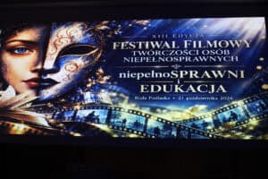 Ekran projekcyjny z grafiką twarzy kobiety i maski weneckiej oraz napisem „XIII Edycja Festiwal Filmowy Twórczości Osób Niepełnosprawnych – niepełnoSPRAWNI i EDUKACJA, Biała Podlaska, 21 października 2026”.