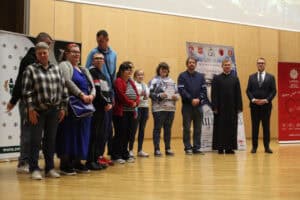 Duża grupa laureatów pozuje na scenie z dyplomami i statuetkami. Obok stoją ksiądz i organizatorzy. W tle plakat festiwalowy i banery reklamowe.