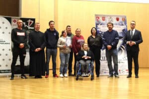 Grupa laureatów i uczestników festiwalu pozuje na scenie z zaproszonymi gośćmi i księdzem. Jedna osoba siedzi na wózku i trzyma statuetkę, a inna trzyma dyplom.
