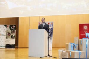 Jakub Semeniuk przemawia za mównicą. W tle jasna ściana z paneli. Po prawej stoją paczki różnej wielkości w brązowym papierze i z błękitnymi wstążkami.