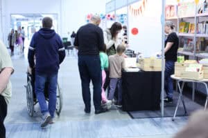 Na hali targowej znajduje się stoisko z książkami, przy którym stoją dorośli i dzieci. Obok przechodzi osoba prowadząca wózek inwalidzki.