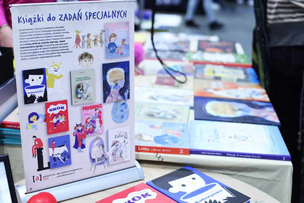 Pionowy plakat reklamowy z napisem „Książki do zadań specjalnych” stoi na stole obok ekspozycji książek dla dzieci.