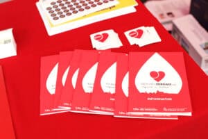 Na czerwonym obrusie leżą czerwone informatory i ulotki akcji „Kierunek Zdrowie – wsparcie w chorobie” oraz małe naklejki z sercami.