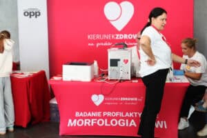 Stanowisko informacyjno-medyczne z czerwonymi banerami akcji „Kierunek Zdrowie – wsparcie w chorobie”. Przy stanowisku stoją organizatorzy i uczestnicy.