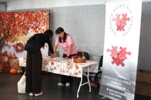 Przy stoisku Fundacji Razem Dla Pokoleń rozmawiają dwie kobiety. Na stoisku znajduje się czerwone logo w kształcie puzzla oraz stolik z obrusem w jesienne wzory.