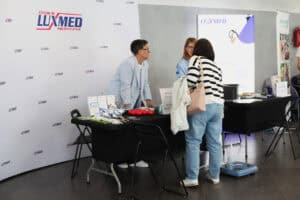 Przy stoisku Centrum Medycznego Luxmed obsługa rozmawia z uczestniczką wydarzenia. Na stoisku leżą materiały profilaktyczne.