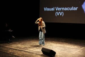 Kobieta gestykuluje na scenie. Na ekranie za nią widnieje napis „Visual Vernacular (VV)”.