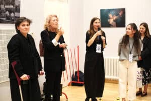 Cztery kobiety stoją w galerii przy ścianie ze zdjęciami teatralnymi. Od lewej: kobieta z kulą ortopedyczną, druga kobieta w czarnym żakiecie wskazuje palcem w prawo, trzecia - Justyna Kazek-Góra mówi do mikrofonu, a czwarta kobieta w szarym swetrze i jasnych spodniach patrzy w ich stronę.