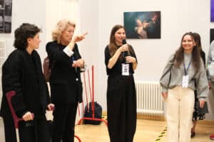 Cztery kobiety stoją w galerii przy ścianie ze zdjęciami teatralnymi. Od lewej: kobieta z kulą ortopedyczną, tłumaczka PJM, trzecia - Justyna Kazek-Góra mówi do mikrofonu, a czwarta kobieta w szarym swetrze i jasnych spodniach uśmiecha się.