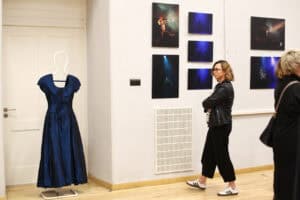 Kobieta w okularach, czarnej skórzanej kurtce i spodniach ogląda zdjęcia na białej ścianie galerii. Obok siebie wisi pięć fotografii z sylwetkami w niebieskim świetle. Przy białych drzwiach stoi wieszak z długą, ciemnoniebieską suknią wieczorową.