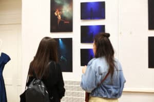 Dwie kobiety oglądają fotografie teatralne na białej ścianie galerii. Na zdjęciach widać postacie w ciemności i niebieskim świetle.