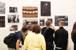 Grupa osób ogląda wystawę fotografii teatralnych eksponowanych na białej ścianie. Wśród zdjęć różnej wielkości są kolorowe fotografie z prób i kulis przedstawień.