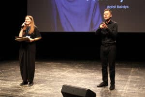 Justyna Kazek-Góra stoi po lewej stronie sceny, trzyma mikrofon i uśmiecha się. Po prawej stoi tłumacz języka migowego. Na ekranie za nimi widnieje napis „Rafał Bołdys”.