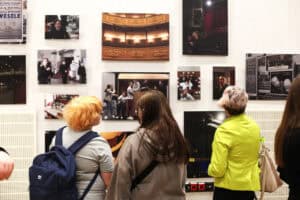 Trzy osoby oglądają fotografie teatralne na białej ścianie galerii. Wśród nich znajduje się duże, kolorowe zdjęcie widowni teatru.