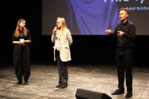 Na scenie stoją trzy osoby. Kobieta w jasnej marynarce i okularach trzyma mikrofon i mówi. Po jej lewej stronie stoi kobieta w czarnym stroju, a po prawej tłumacz języka migowego.