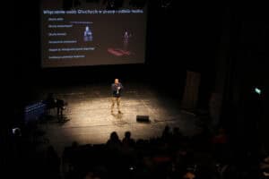 Prelegent stoi na scenie w sali teatralnej. Na ekranie za nim widnieje tytuł „Włączenie osób Głuchych w pracę i odbiór teatru” oraz lista punktów. Na widowni siedzi publiczność.
