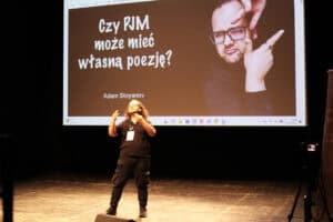 Adam Stoyanov, mężczyzna w okularach i z długimi włosami, gestykuluje na scenie. Na ekranie za nim widnieją tytuł „Czy PJM może mieć własną poezję?” i jego imię oraz nazwisko. W prawym górnym rogu ekranu znajduje się jego portret.