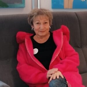Grażyna Chaberko w amarantowej bluzie, na tle szarego oparcia kanapy.