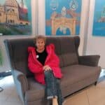 Grażyna Chaberko siedzi na szarej kanapie, ubrana w amarantową bluzę. Na ścianach za nią wiszą obrazy z motywami architektonicznymi i sakralnymi.